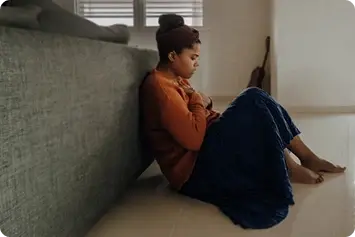 A woman sitting on the floor with her back against the bed looking sad