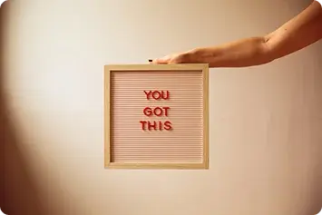 A wooden board that reads You Got This with a hand resting on it